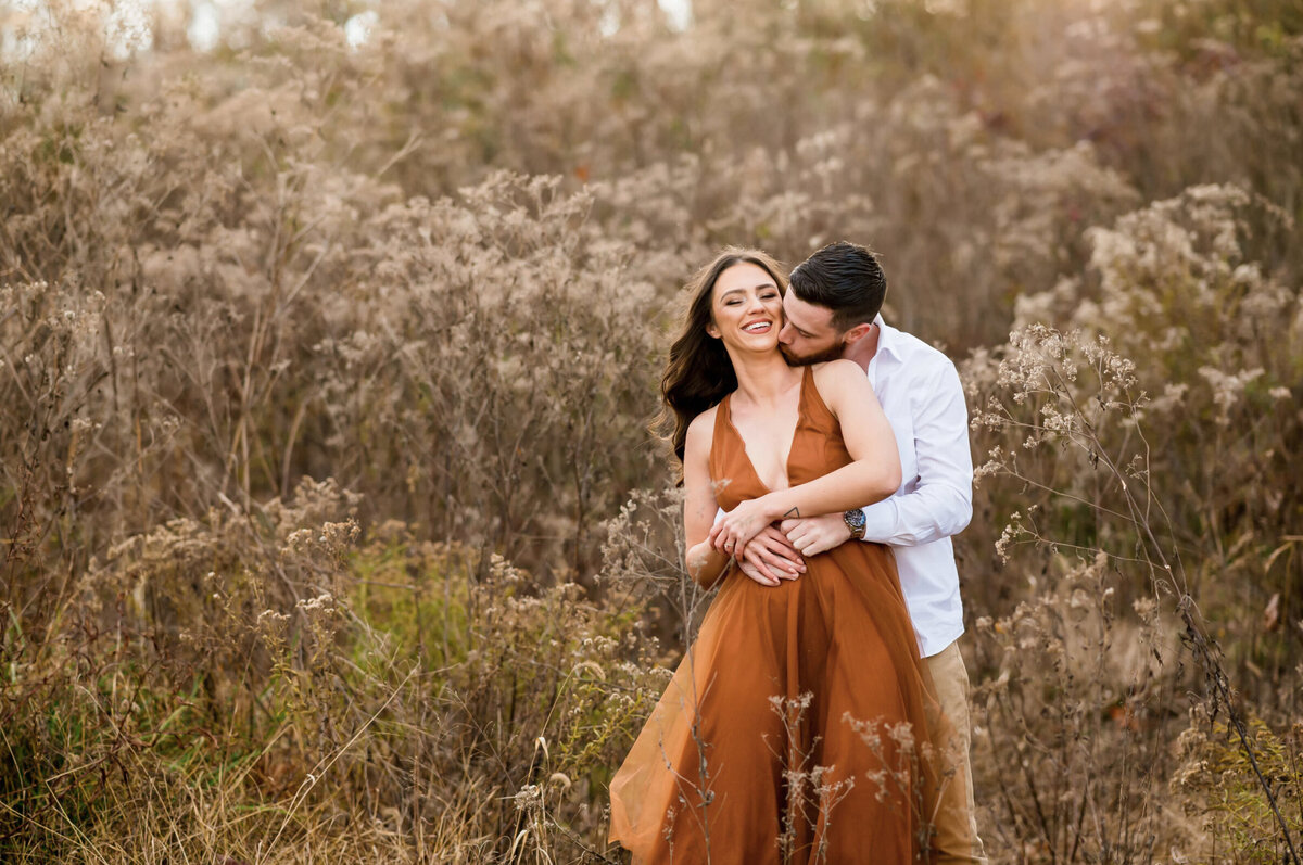 Woman wearing a brown dress, and a man wearing a white shirt standing in a field posing for their engagement shoot