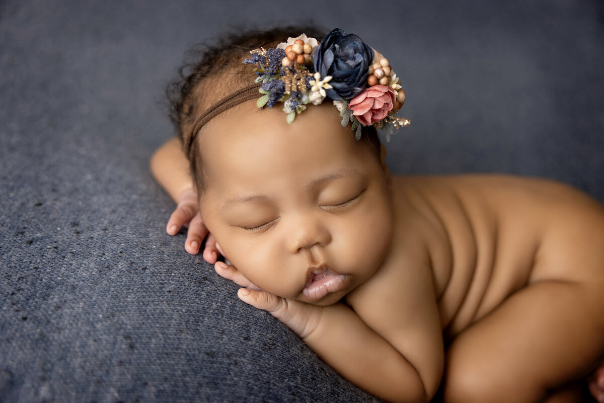 A sleeping baby rests on a dark gray blanket, lying on their side with hands under their cheek. Adorned with a floral headband in navy blue, pink, and peach, this serene scene is captured beautifully by a Jacksonville newborn photographer, conveying warmth and tranquility.