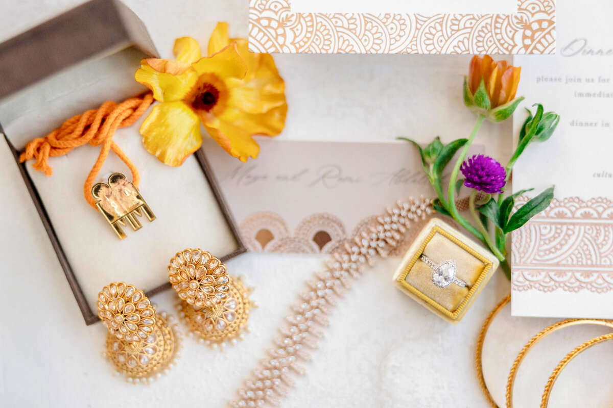 A flat lay of traditional wedding items featuring a pair of gold earrings, a gemstone necklace, a yellow flower, and two invitation cards. A gold ring is displayed in a velvet box, with colorful flowers adding decorative touches.