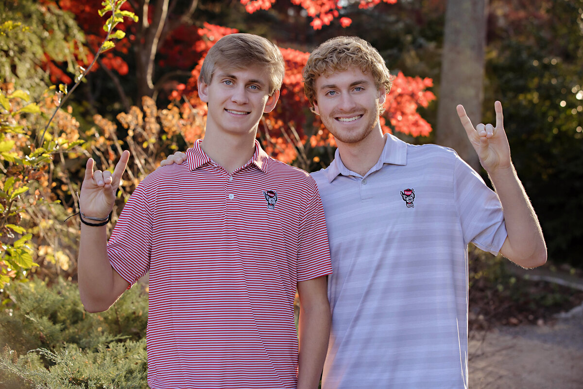 Lively fall image of college senior and freshman brothers in stylish NC State fan outfits standing together in a leafy garden at UNCC Botanical Garden