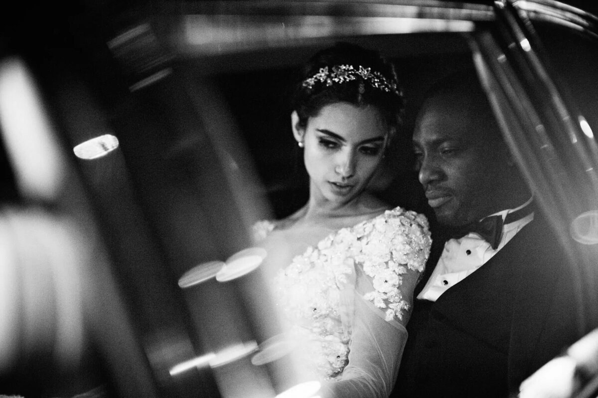A couple shares an intimate moment in the backseat of a car, the bride's wedding dress and the groom's tuxedo elegantly illuminated by the light.