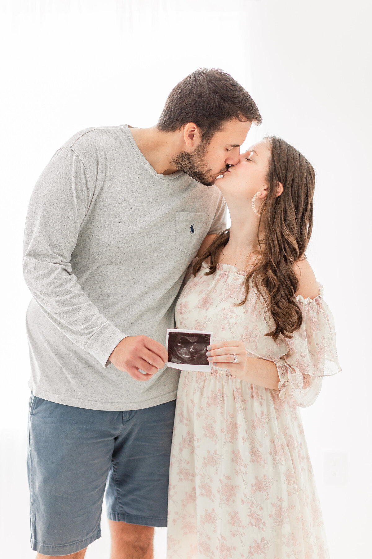 A pregnancy announcement photo of a couple kissing while holding a sonogram in front of a window by dc maternity photographer