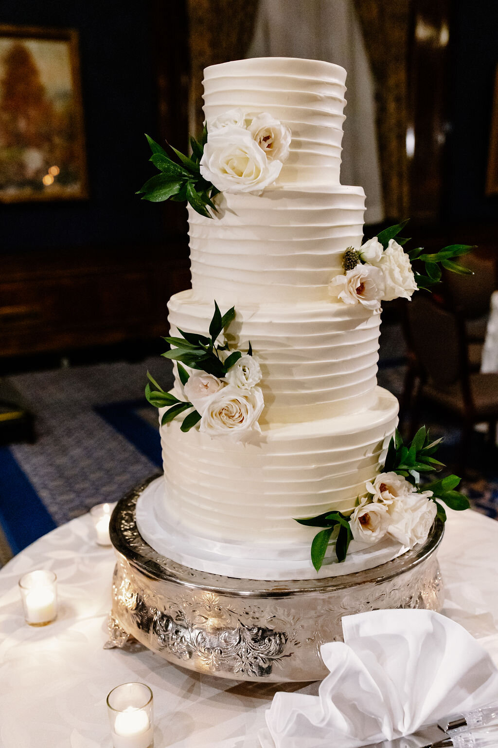 hA four-tier white wedding cake decorated with delicate white roses and green leaves sits on table at wedding reception in Chicago.