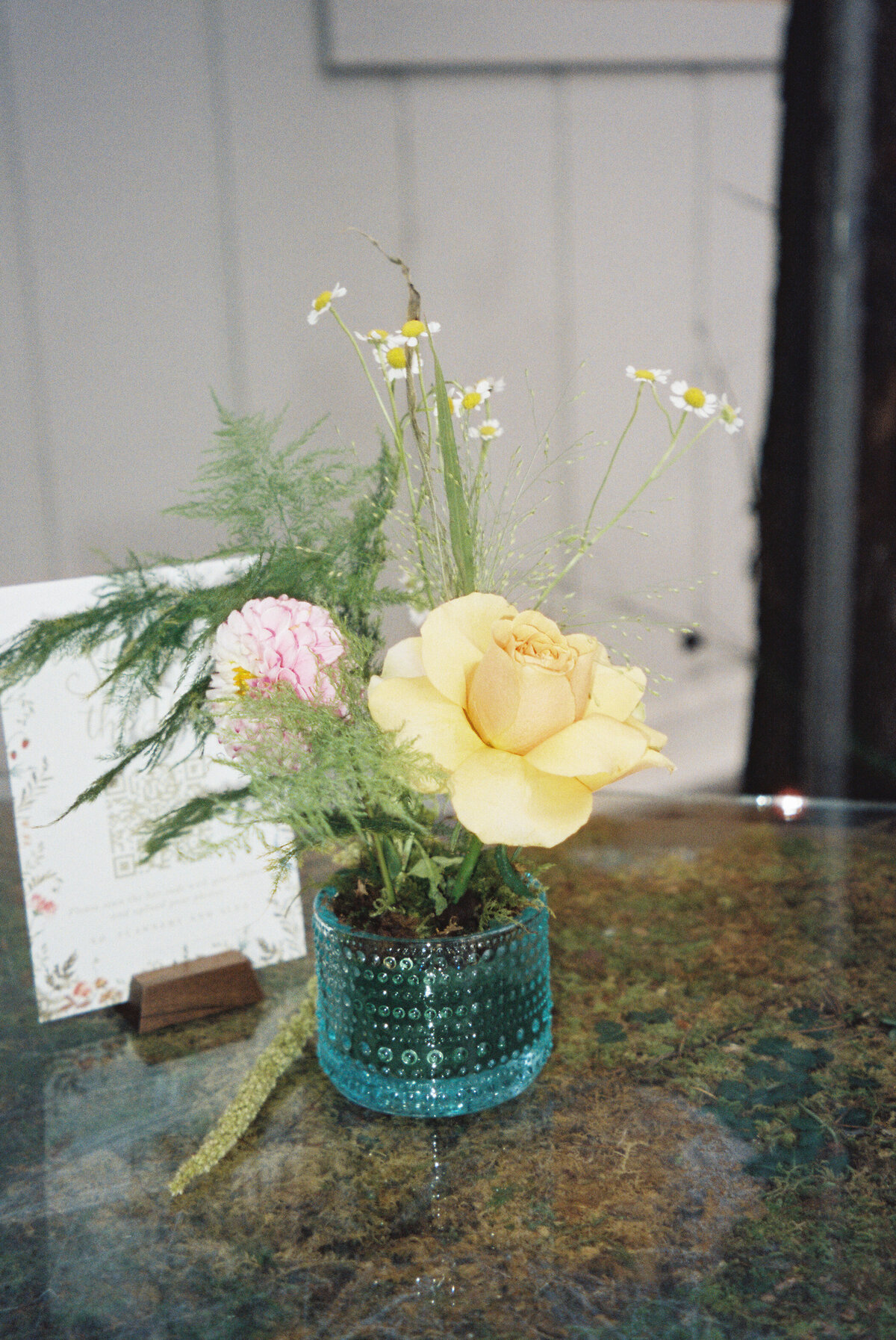 Small garden-inspired floral arrangement with a yellow garden rose, chamomile, delicate greenery, and a pink dahlia displayed in a textured blue vase—created by a Northwest Arkansas wedding florist for an intimate reception detail.