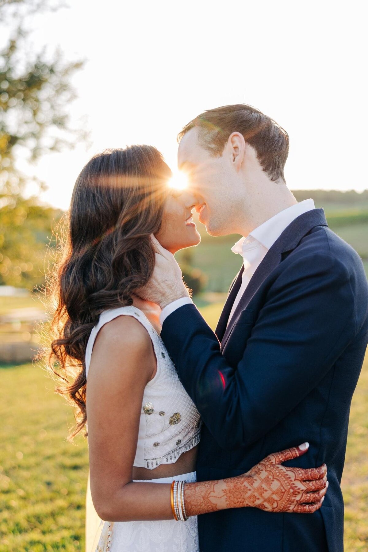 A couple in wedding attire embracing and touching foreheads lovingly in a sunlit field at sunset.