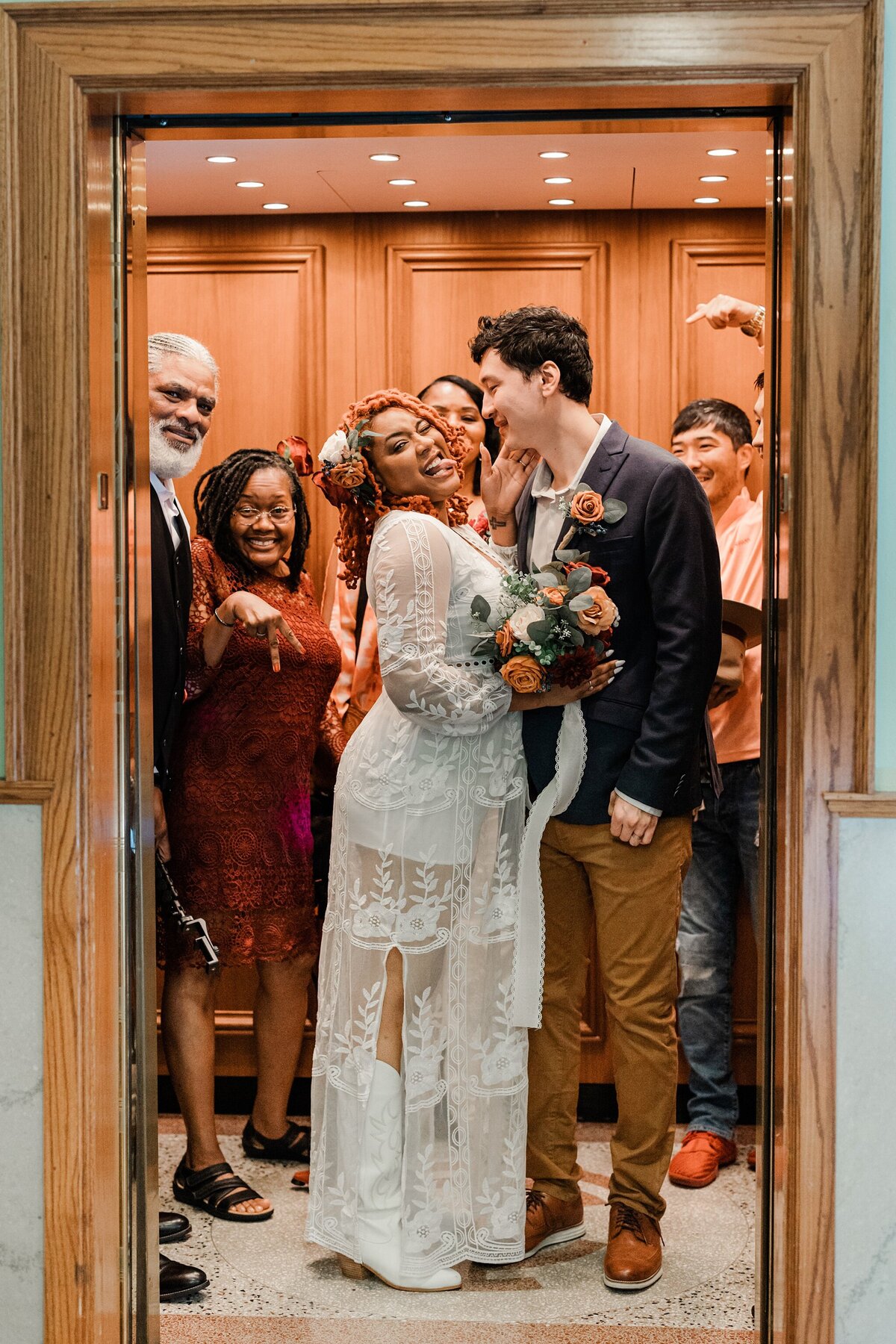 A bride, groom, and their families all playfully posing together on the elevator at the Tarrant County Courthouse in Fort Worth, Texas. The bride is on the left and is wearing an intricately patterned, white dress with white cowboy boots, and a white hair piece. She playfully smiles and sticks out her tongue while holding a bouquet. The groom is on the right and is wearing a dark blue jacket and brown dress pants. He looks like he's about to lean in to kiss the bride on the cheek. Their families form a semicircle around them, and they are all laughing, joking, and celebrating.