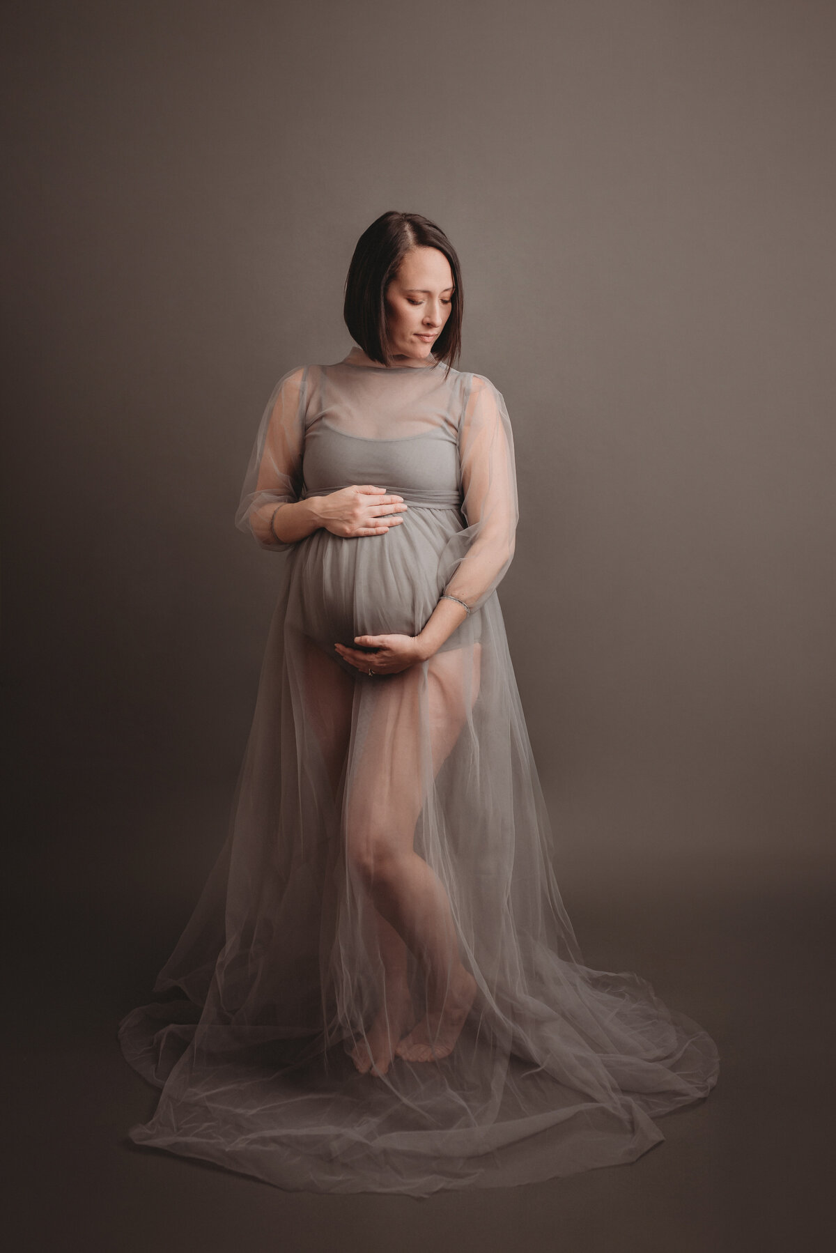 Pregnant woman posing for portraits standing wearing grey tulle dress holding baby bump