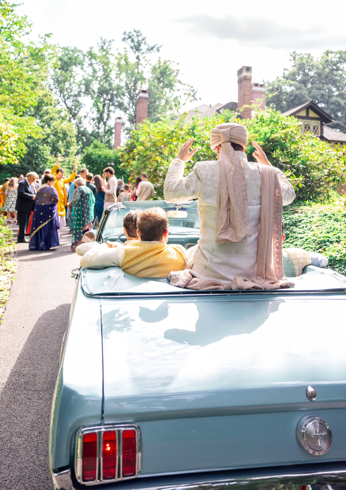 A joyful couple in traditional attire sits on the back of a light blue convertible. They are surrounded by greenery and sunlight, with a group of people gathered in the background, suggesting a celebratory event.
