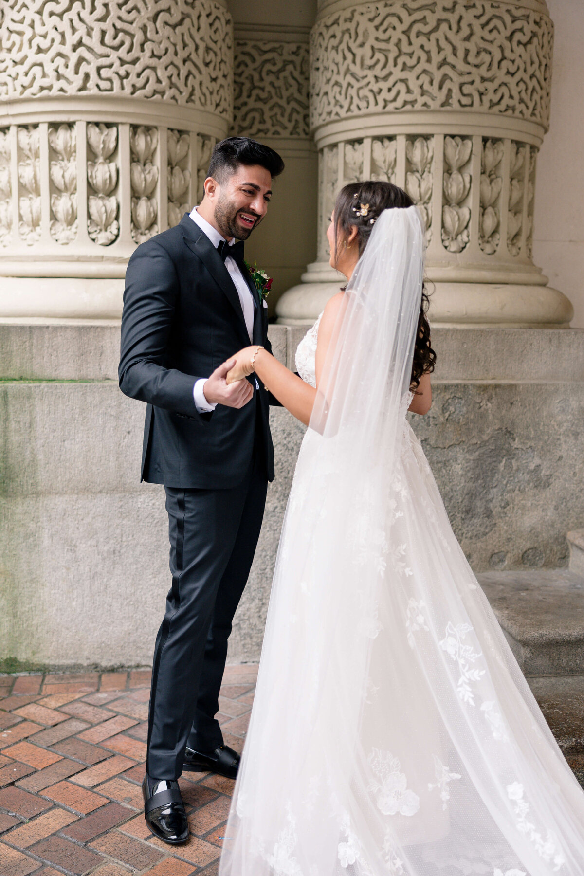 A bride and groom smile at each other while holding hands in front of an ornate stone building. The bride wears a white gown and veil, and the groom is in a black suit with a bow tie.