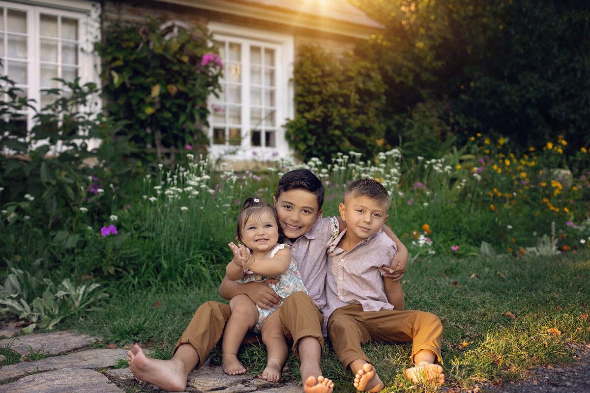 A young boy sits in a lawn full of wildflowers hugging onto his toddler little brother and sister taken by a New Jersey Family Photographer