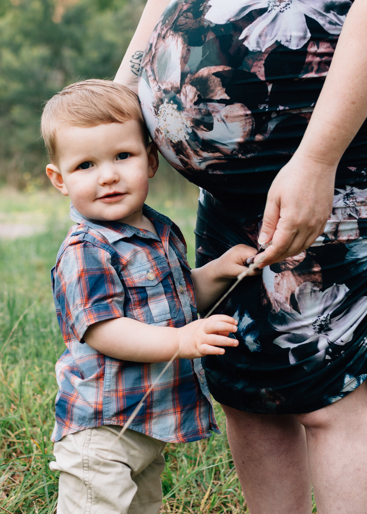 Young son looking at  camera, standing next to mother's belly during outdoor maternity session.
