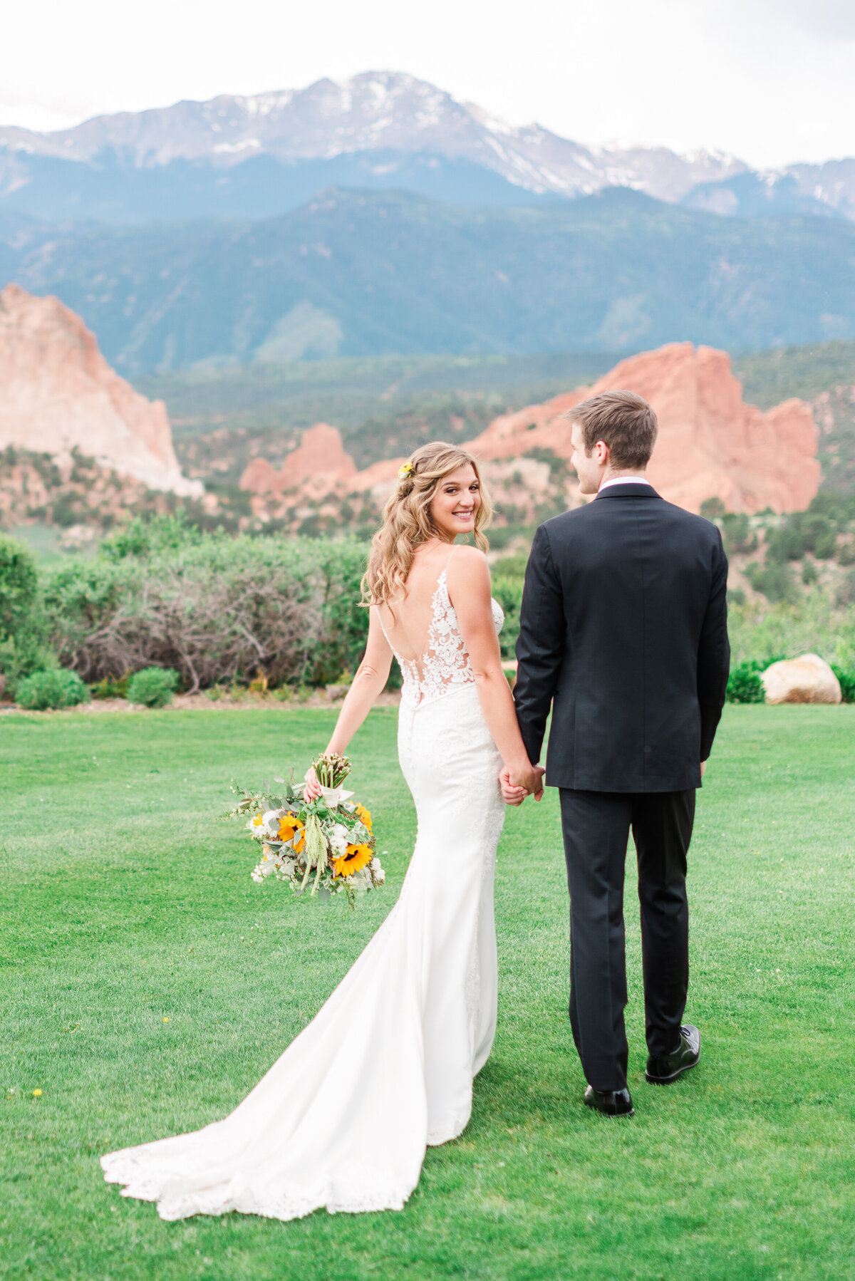 Bride smiling over her shoulder while holding hands with her husband, on a green lawn overlooking Garden of the Gods.