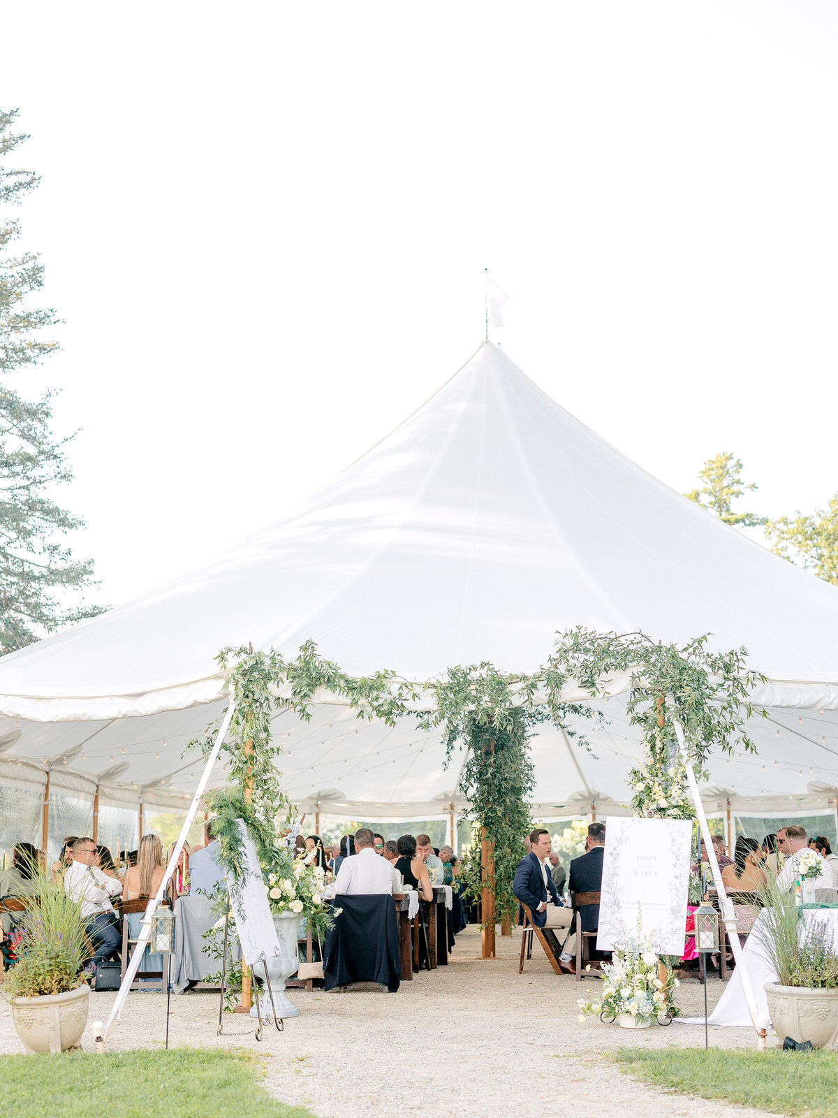Outdoor shot of wedding reception tent with greenery draped on it and wedding guests at tables underneath