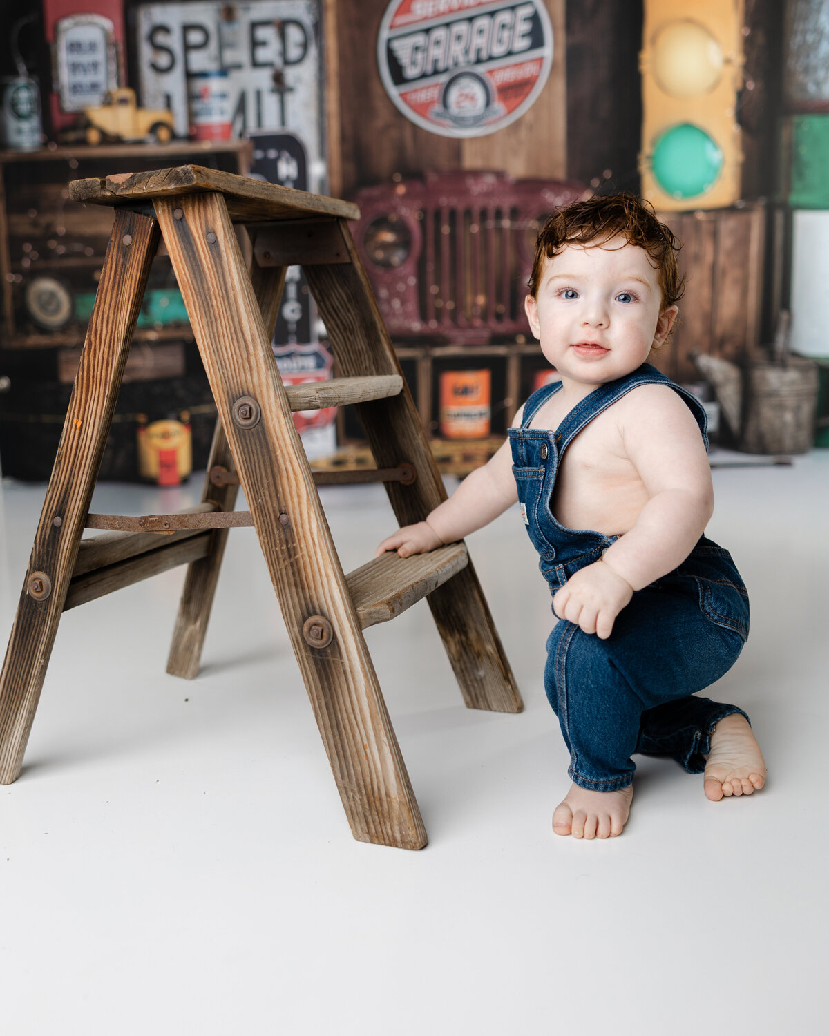 Happy one-year-old boy posing with a small wooden ladder in front of a vintage truck-themed cake smash set. Photograph by Yucaipa cake smash photographer Kaitlyn Dawn Photography.
