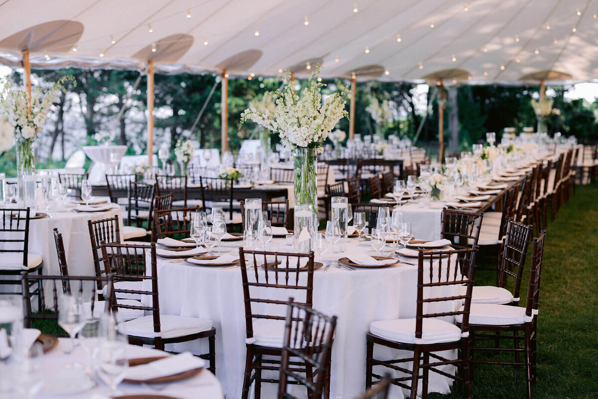The Cape Cod Summer Tent is set up with elegant tables and chairs, white table cloths, white flower centerpieces, and cutleries.