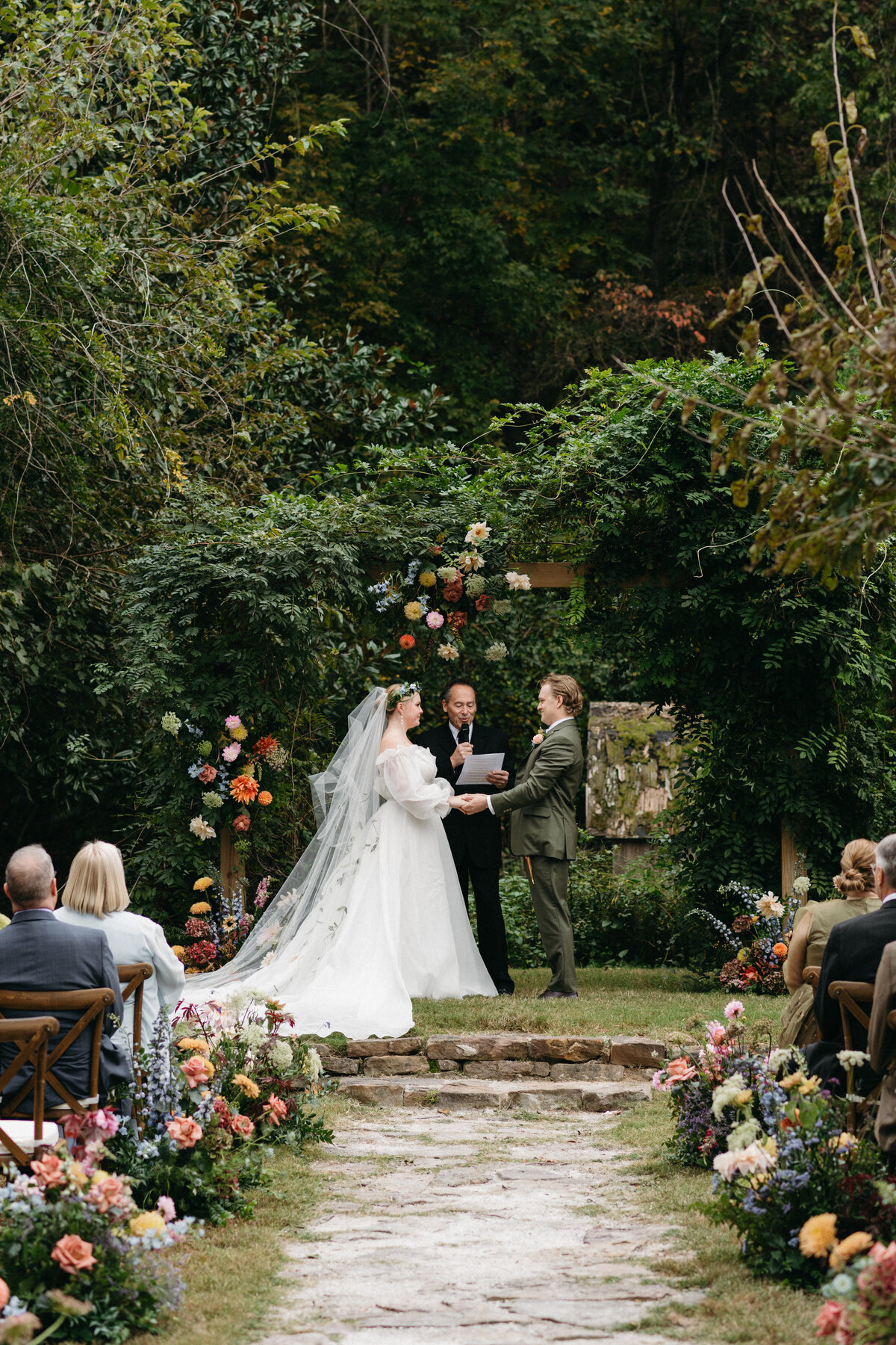 Bride and groom exchanging vows beneath a lush green ceremony arch overflowing with colorful dahlias, roses, and wildflower-inspired blooms, surrounded by whimsical aisle arrangements for a nature-inspired outdoor wedding in Northwest Arkansas, designed by a fine art wedding florist.