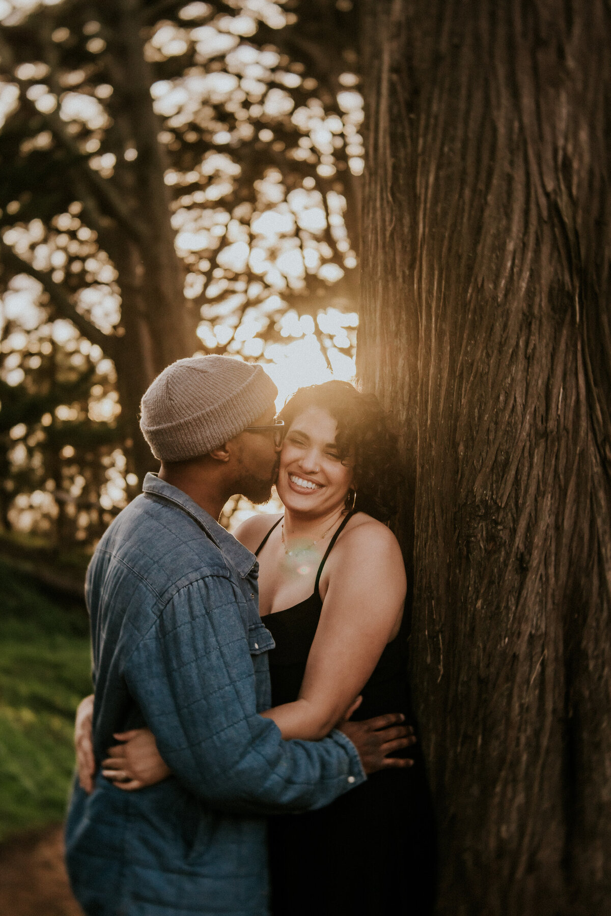 Bay Area couple leans against tree and man kisses women's cheek.  She wears black dress and he is in a jean shirt and beanie