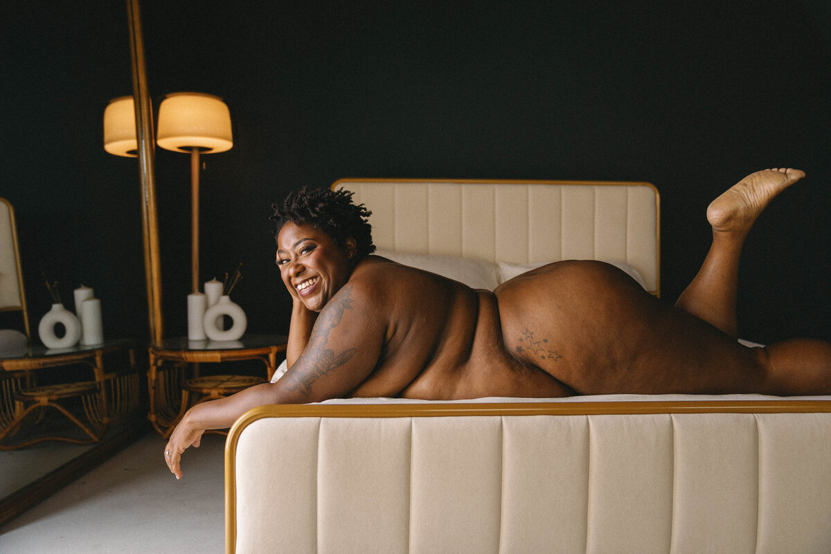 A nude client lays on their stomach across a bed smiling with their feet up in the Good Bodies studio