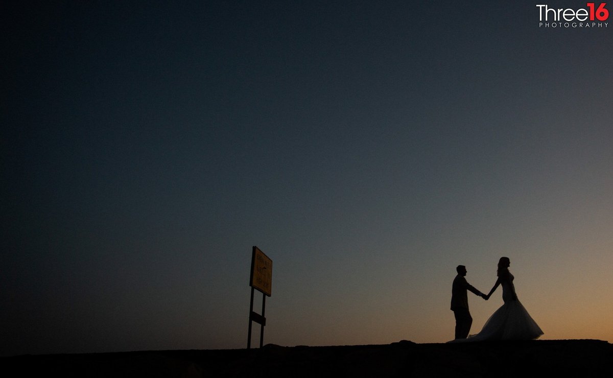 Bride and Groom walk along the beach holding hands just before the sun goes down leaving them as a silhouette