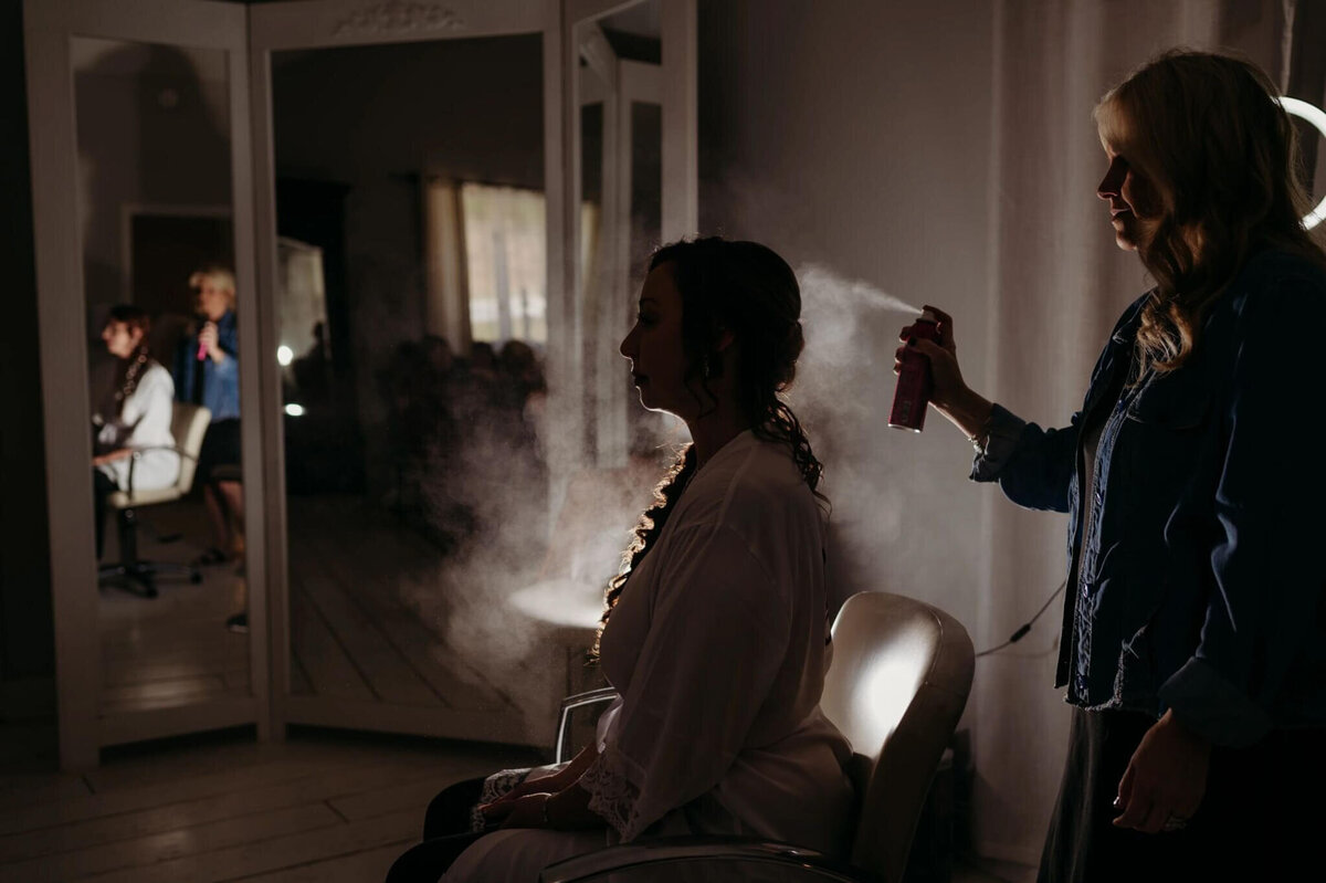 Photo of a silhouette of a bride and a white robe getting hairspray in her hair