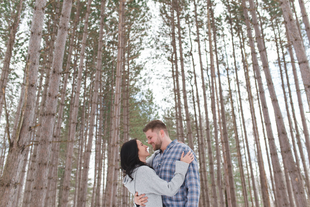 engaged couple standing together among tall pine trees