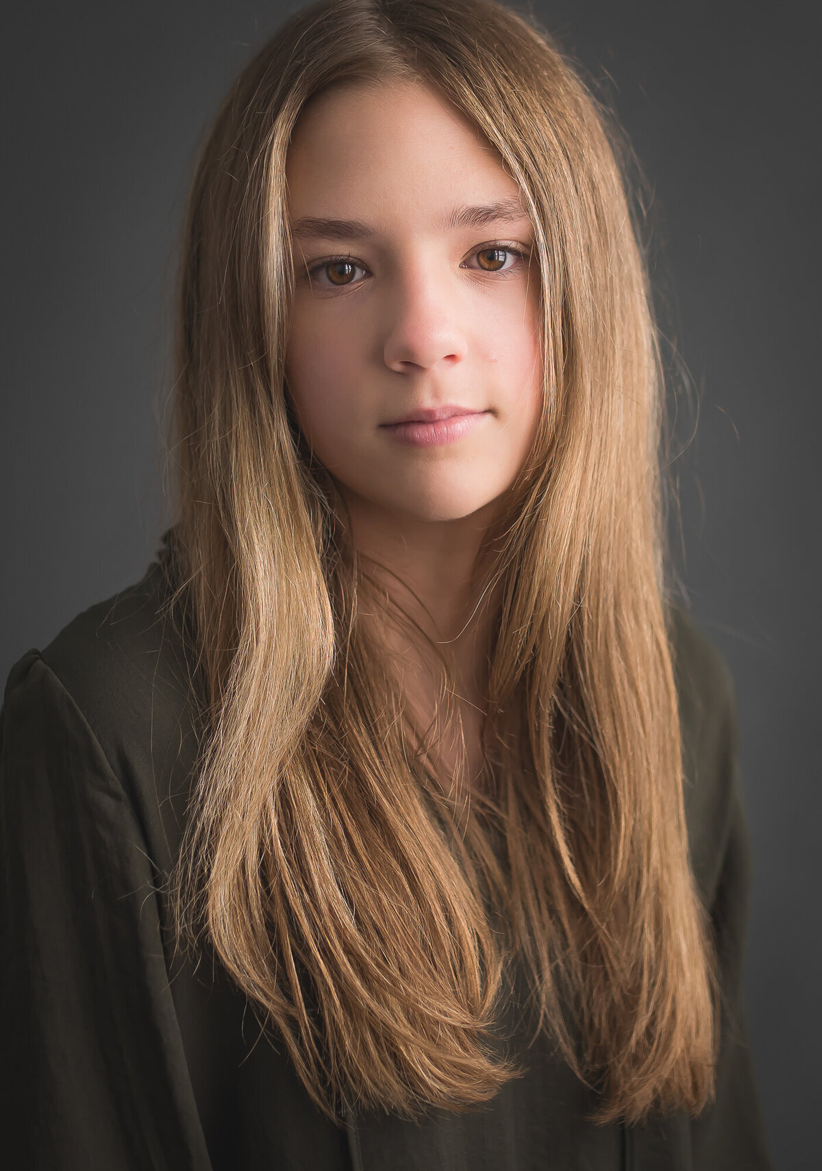color portrait of a senor girl in a dark olive blouse with long blonde hair with a grey background