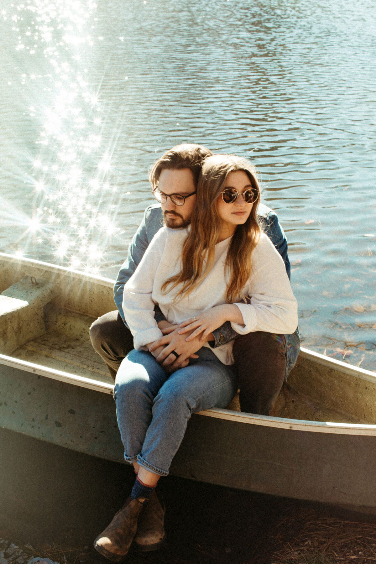 A guy is sitting in a canoe wrapping his arms around a girl with her legs hanging over the side of the boat as the sun reflects off the water behind them.