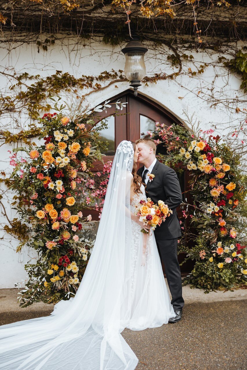 Newlyweds kiss while standing in front of a large floral display with a matching bouquet