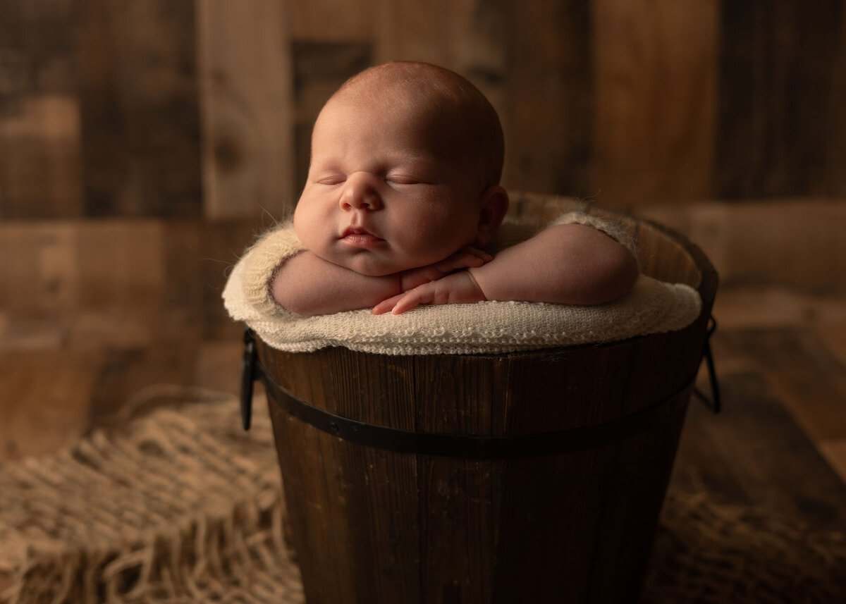 Maddie Rae Photography baby is in a dark wooden bucket. he is sleeping and has his head resting on his hands, they are on the edge of the bucket