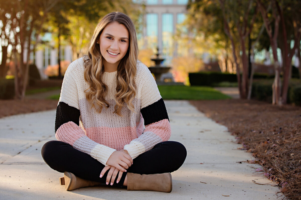 Happy High school senior during senior portrait session on a Fall afternoon in Ballantyne.  Senior is sitting criss cross applesauce on the concrete in a garden with a fountain in the backdrop.