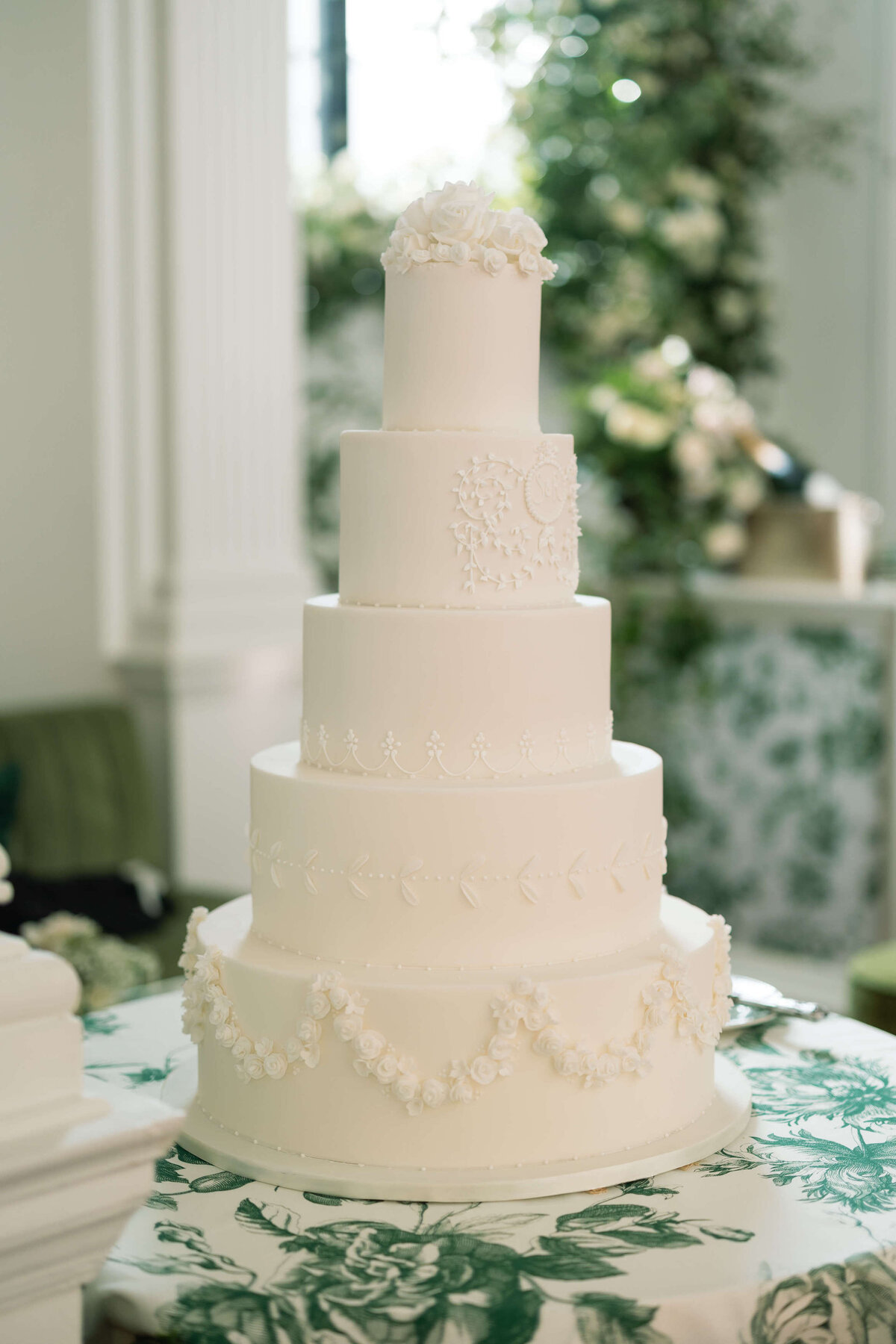 white five tier wedding cake with intricate piping decoration on a green and white patterned tablecloth with a matching bar behind it for a chic wedding