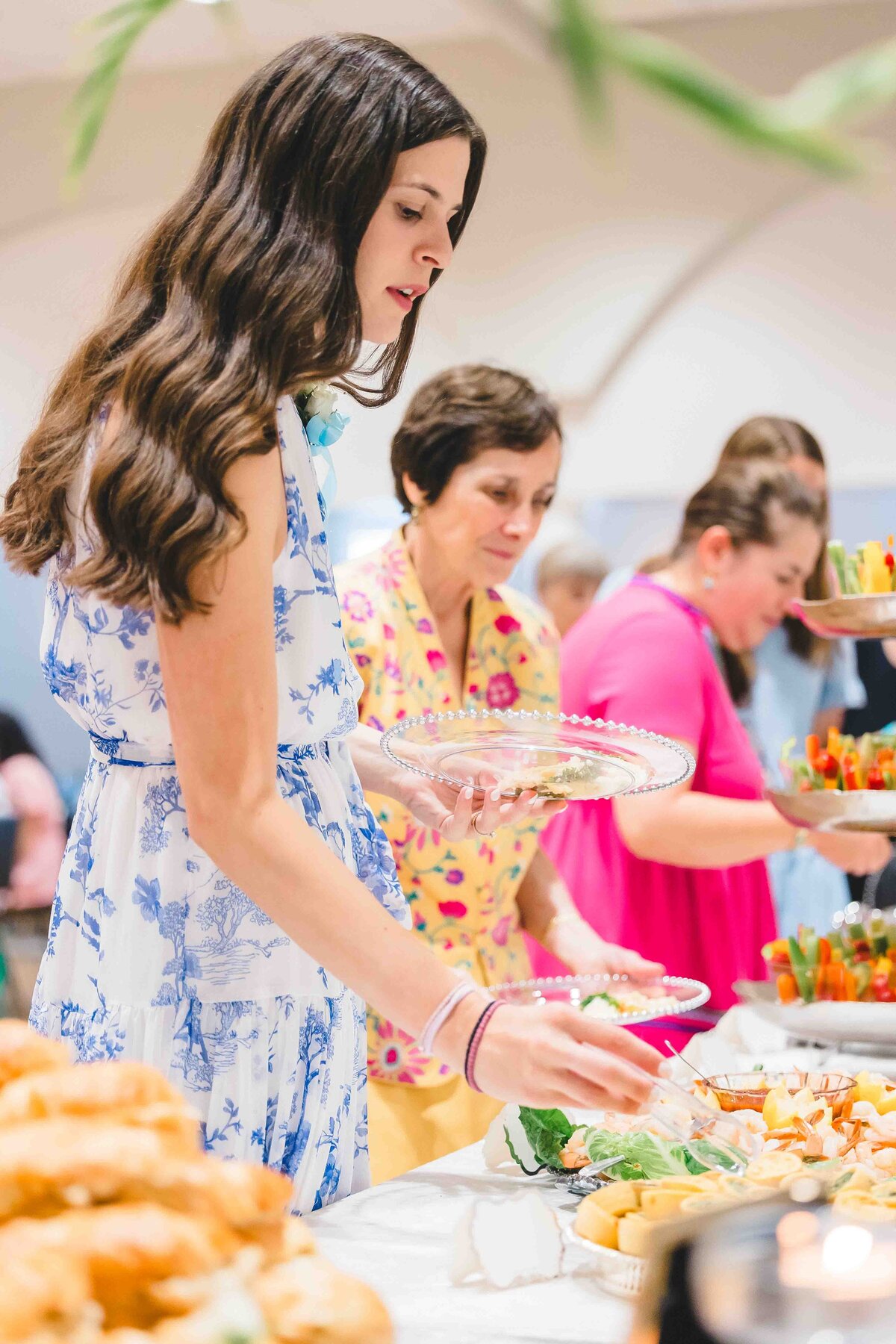 A group of people serves themselves food from a buffet, captured by an event photographer in Fayetteville, NC. The nearest person, in a blue and white dress, holds a clear plate. The table brims with dishes like sandwiches and vegetables at this inviting social gathering.