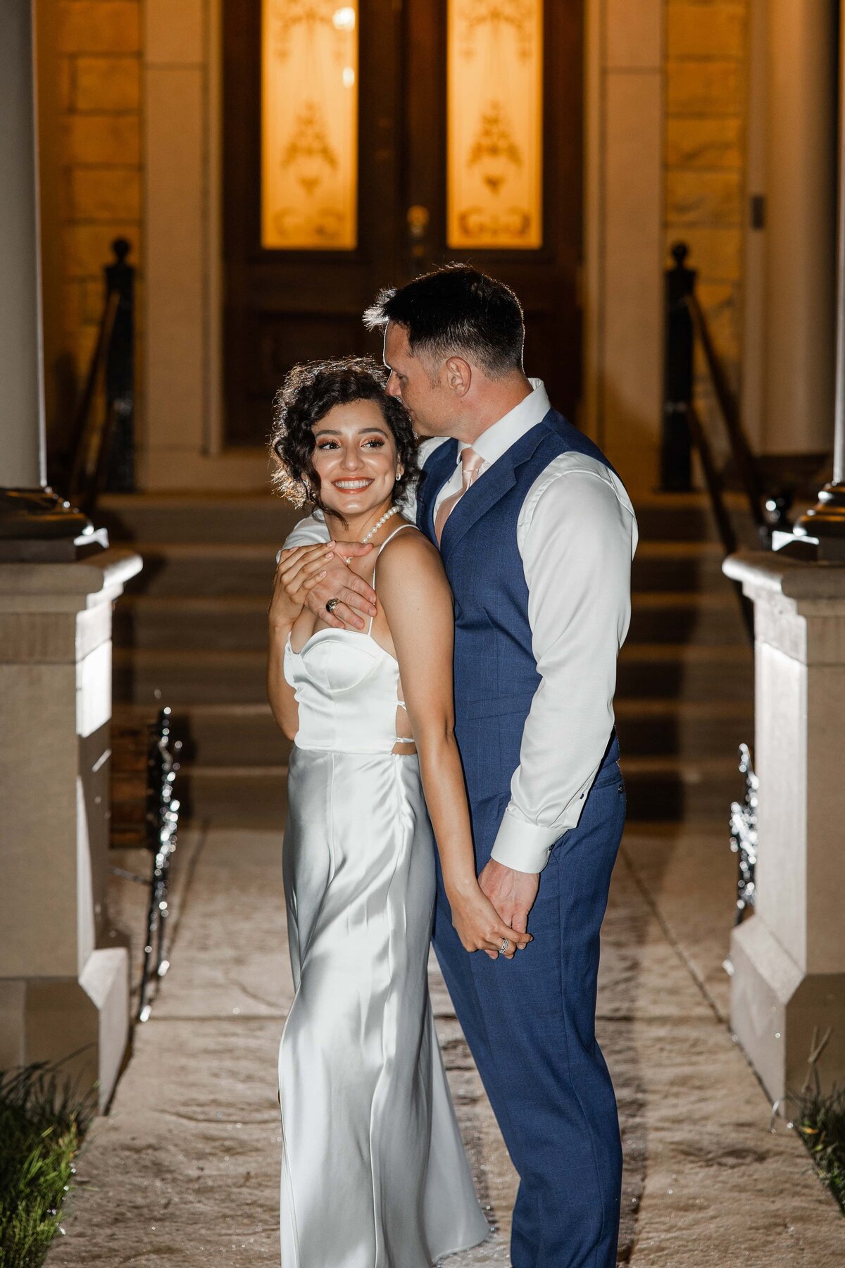 A bride in a white satin dress and a groom in a blue suit embrace lovingly outside a grand building at night during their park farm winery wedding.