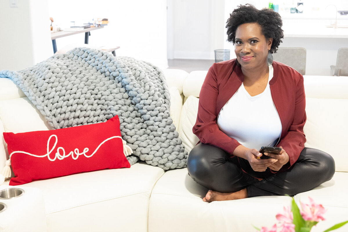 An image of a woman sitting on a white coach with her legs on in and crossed next to a red pillow that says love