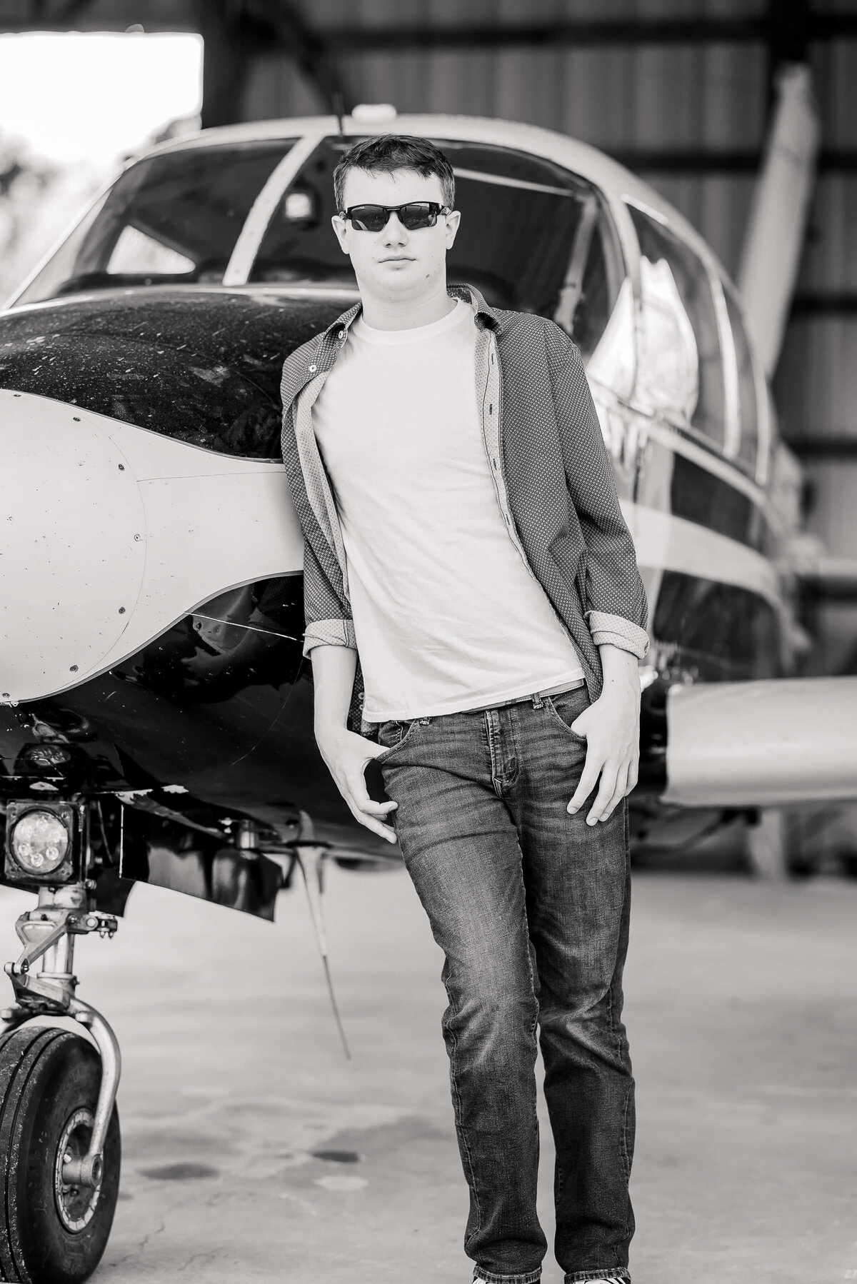 A person wearing sunglasses leans against the front of a small airplane inside a hangar. They are dressed in a casual outfit with a t-shirt, open button-down shirt, and jeans. The image is in black and white.