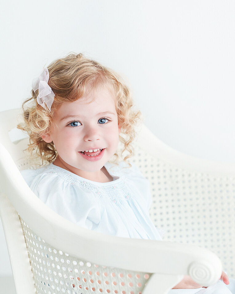 3 year old girl photographed in a white background studio in st simons sialnd wearing a feltman brothers smocked dress in a white wicker chair