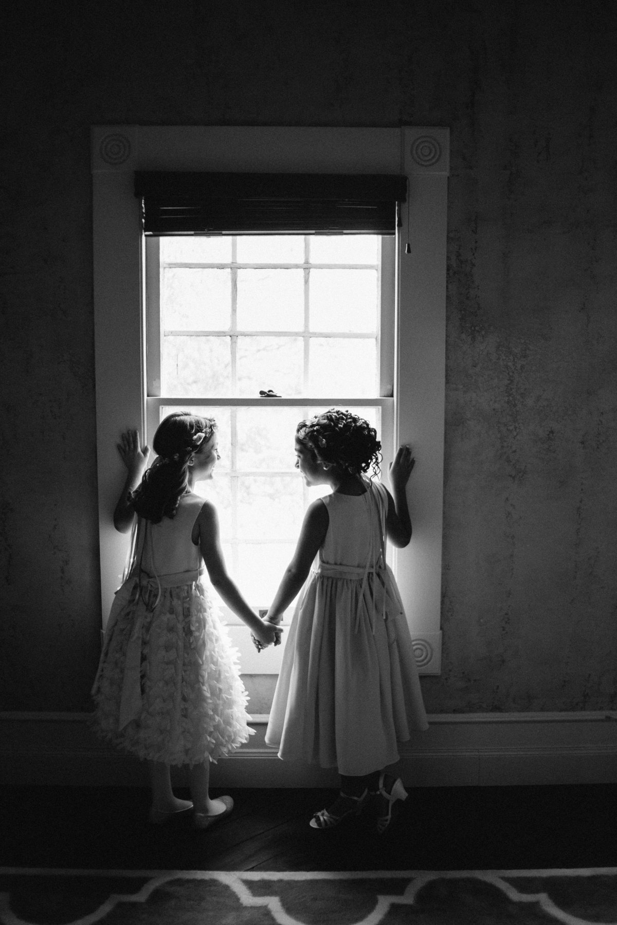 flower girls holding hands and looking out window while getting ready before ceremony at The Grande Hall at Hoffman Ranch