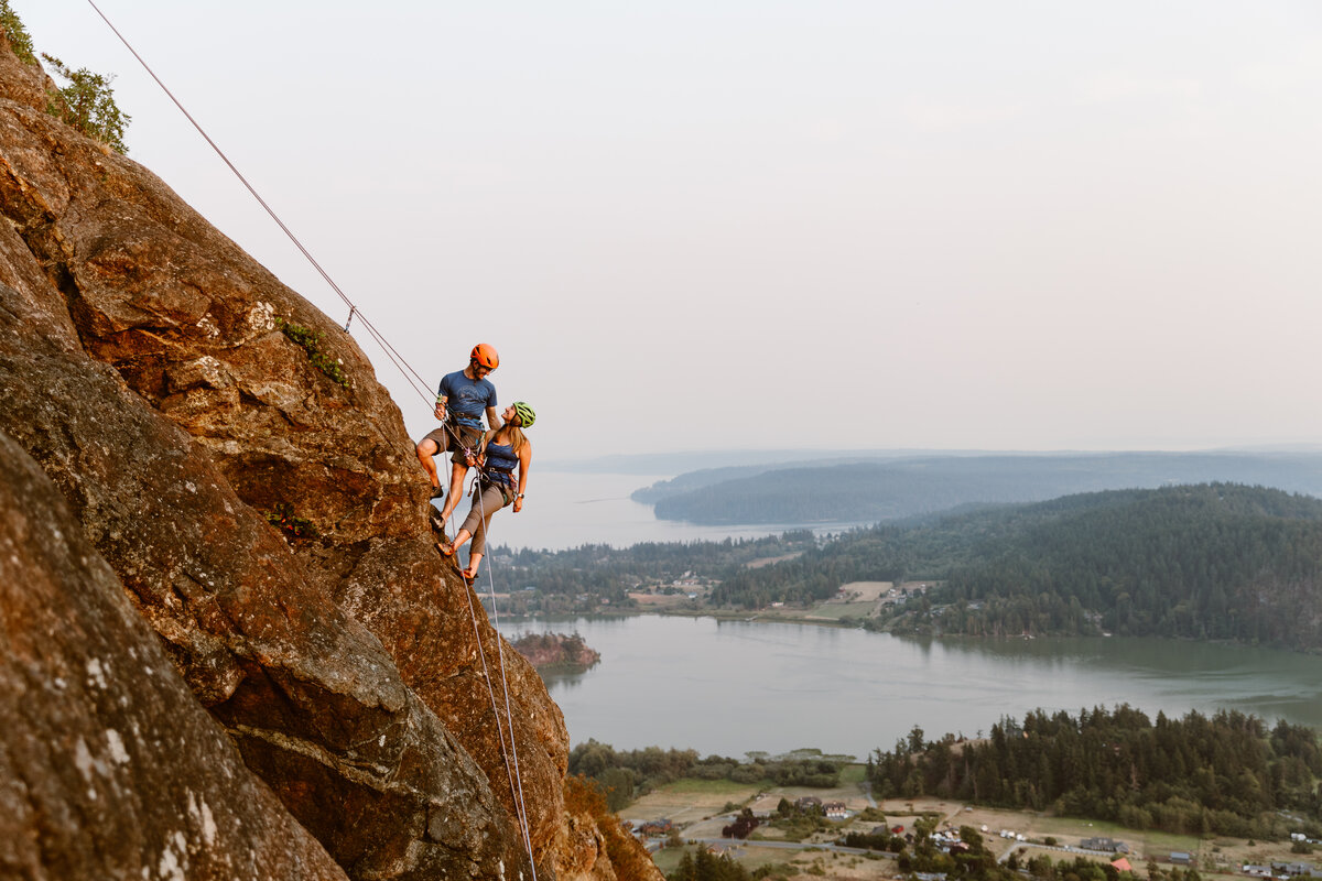 couple on rope with harnesses and helmets rappelling down cliff during rock climbing engagement session