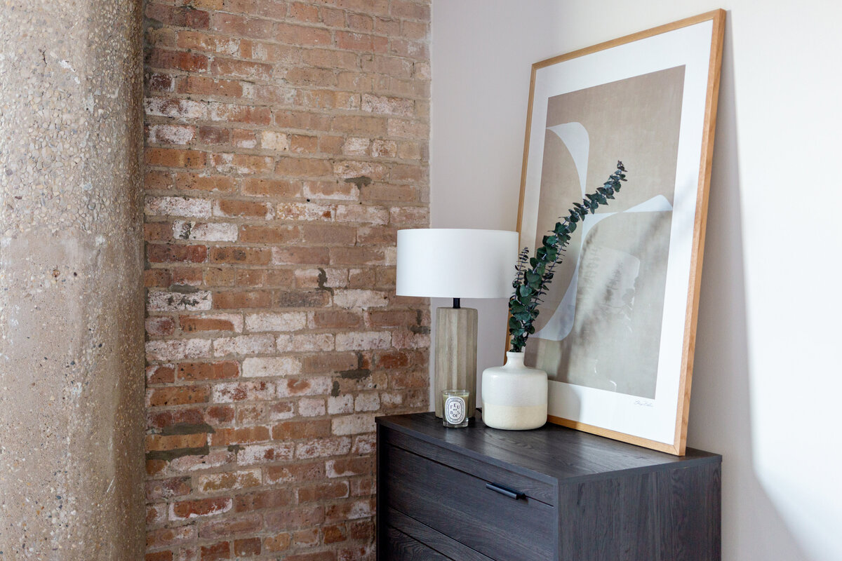 Brick wall next to nlack wooden dresser with scalloped concrete table lamp on top, white ceramic vase filled with eucalyptus and diptyque candle and abstract beige wooden framed artwork