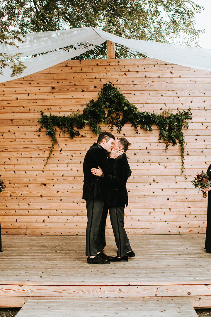 Two grooms share a kiss during their wedding on a wooden altar, with green foliage in the background.