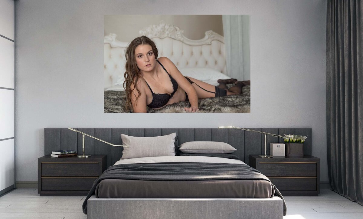 A canvas that is hung over a bed. The photo is of a  brunette woman wearing black lingerie and she is laying on her side looking at the camera.