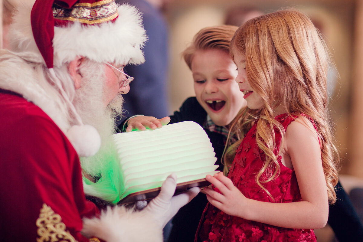 Santa came to visit this family at home, and is showing them his magic book that lights up. They are clearly in awe.