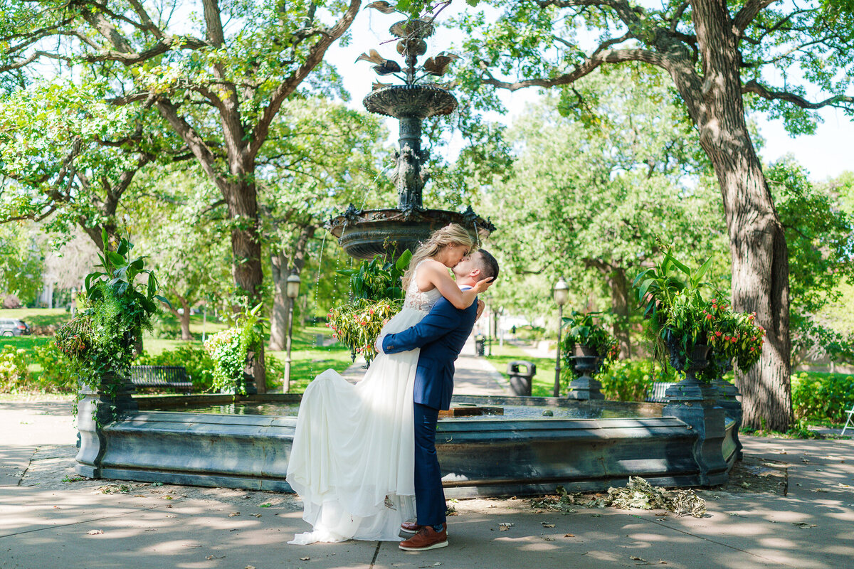 Groom lifts and kisses bride in front of a fountain in Saint Paul, Minnesota.