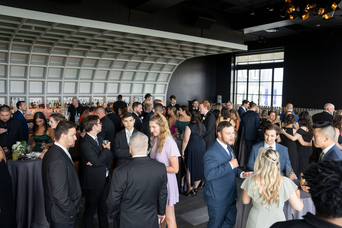 A large group of people in formal attire mingling at a social event in a modern venue with a curved ceiling. People are engaged in conversations, and a few are holding drinks. Tables with floral arrangements are scattered throughout the room.