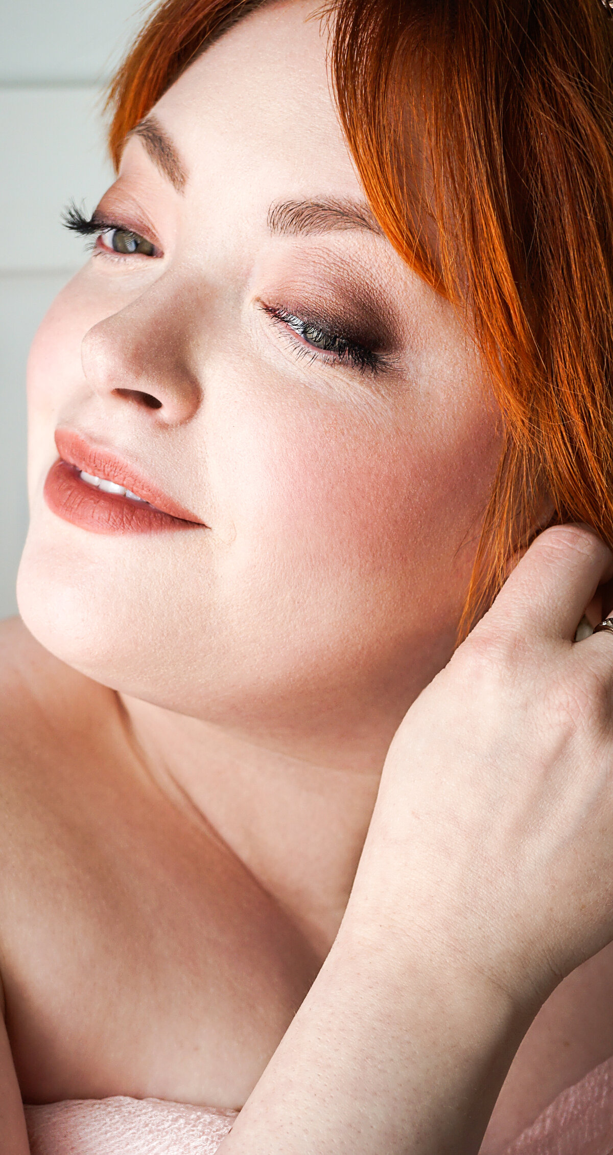 A redheaded bride places an earring in her ear while wearing soft, natural makeup and a smile.