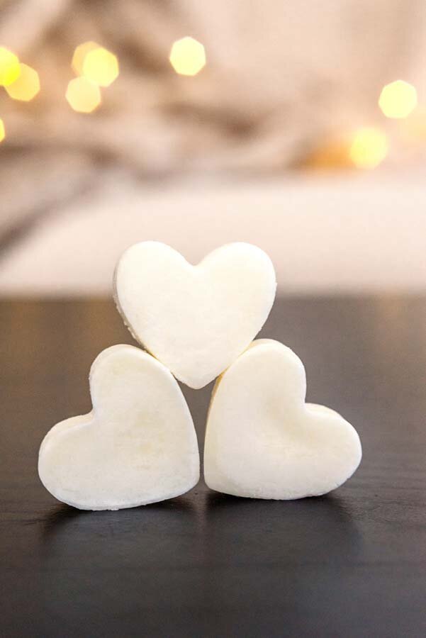 Three heart-shaped wax melts arranged standing on a dark surface, with softly blurred lights in the background.