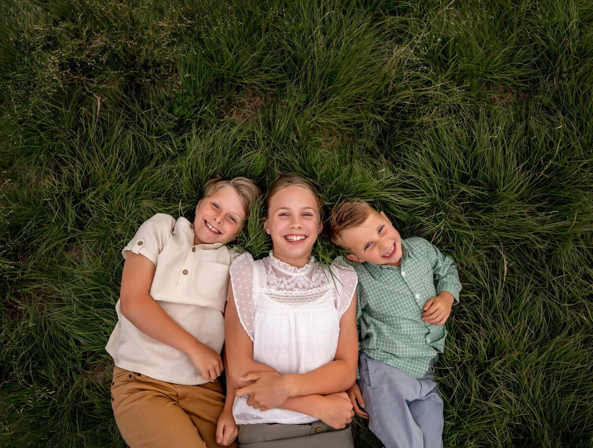 Three siblings lay in the grass and smile for the camera during their portrait session with an Asheville Family Photographer