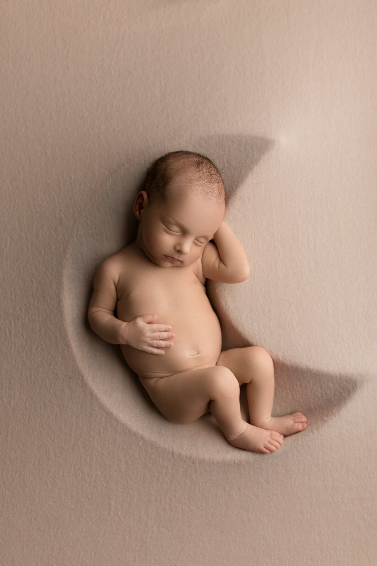 Newborn baby boy is sleeping atop of a moon shape. One of baby's hands is resting on his belly, the other underneath his head. Baby's legs are curled inside of the moon-shape. Image captured by New Jersey's best newborn photographer, Katie Marshall,.