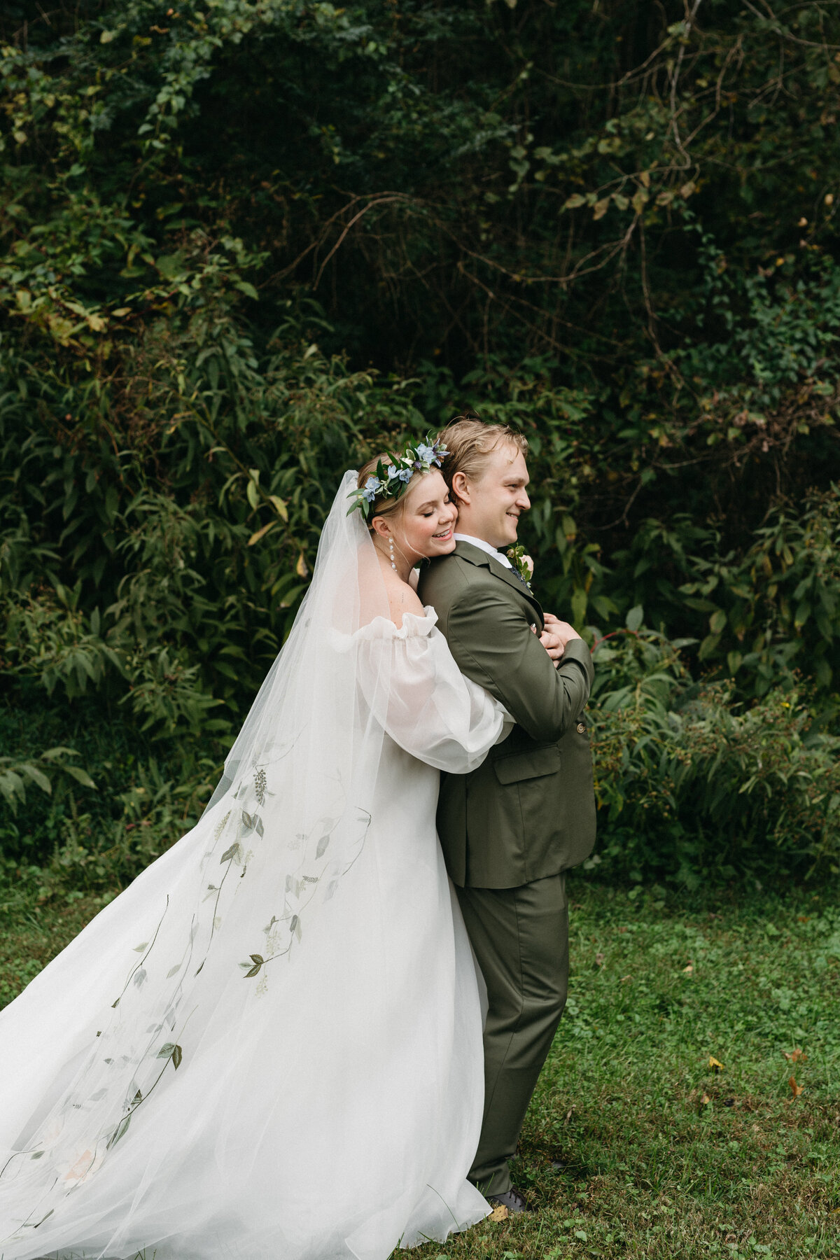 Bride embracing the groom from behind in a wooded outdoor setting, wearing an airy off-the-shoulder gown, floral crown, and long veil embroidered with greenery. Groom wears an olive suit as they pose surrounded by lush natural foliage during their garden-style wedding.