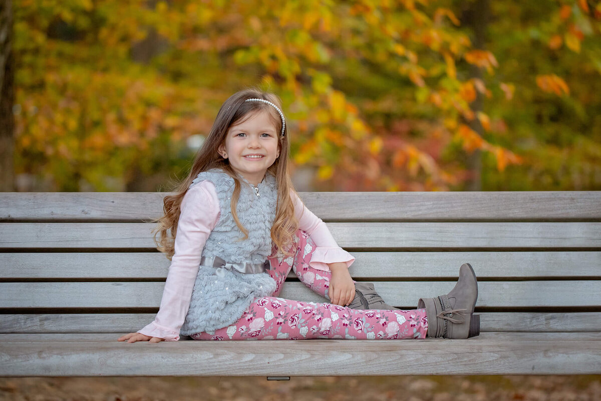 My daughter posing on a bench at the park with fall foliage behind her.